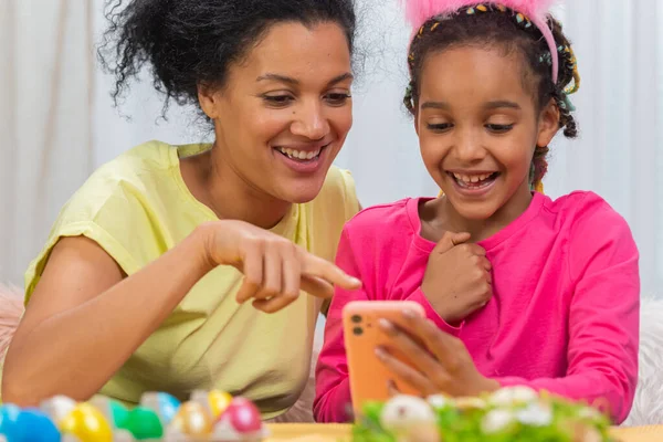 Mamma e figlia con divertenti orecchie da coniglio si scattano selfie e si divertono. Donna afroamericana e bambina sono seduti a tavola in una stanza decorata a festa a casa. Buona Pasqua. Da vicino.. — Foto Stock