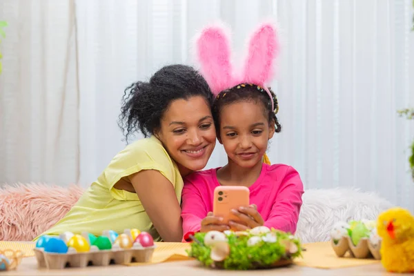 Mamma e figlia con divertenti orecchie da coniglio si scattano selfie e si divertono. Donna afroamericana e bambina sono seduti a tavola in una stanza decorata a festa a casa. Buona Pasqua. Da vicino.. — Foto Stock