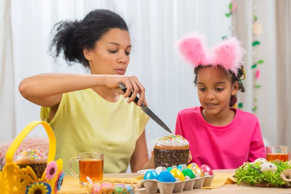 Mamma taglia una torta di Pasqua per curare sua figlia. Donna afroamericana e bambina sono seduti a tavola in una stanza decorata a festa a casa. Buona Pasqua. Da vicino.. — Foto Stock