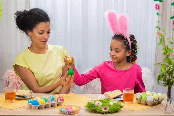 Mamma e figlia si divertono e rompono le uova di Pasqua. Donna afroamericana e bambina sono seduti a tavola in una stanza decorata a festa a casa. Buona Pasqua. Da vicino.. — Foto Stock