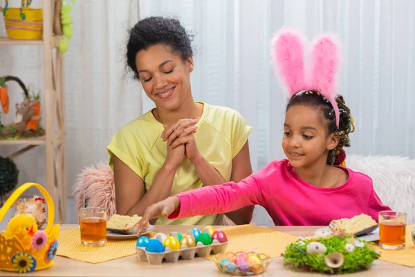 Mamá e hija se divierten y rompen los huevos de Pascua. Mujer afroamericana y niña están sentadas a la mesa en una habitación festivamente decorada en casa. Feliz Pascua. De cerca.. — Foto de Stock