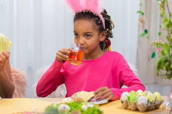 Mamma e figlia con divertenti orecchie da coniglio stanno bevendo il tè con la torta di Pasqua. Donna afroamericana e bambina sono seduti a tavola in una stanza decorata a festa a casa. Buona Pasqua. Da vicino.. — Foto Stock