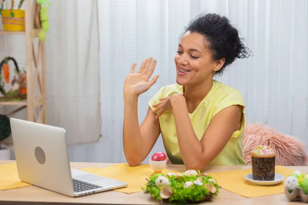 Carina donna afroamericana che parla tramite videochiamata su un computer portatile. Una giovane donna mostra un uovo dipinto e una torta di Pasqua mentre siede a un tavolo da vacanza decorato. Buona Pasqua. Da vicino.. — Foto Stock