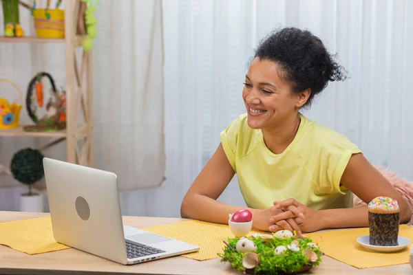 ラップトップでビデオ通話で話すかわいいアフリカ系アメリカ人女性。若い女性は、装飾された休日のテーブルに座っている間に描かれた卵とイースターケーキを示しています。ハッピー・イースター。閉じろ!. — ストック写真