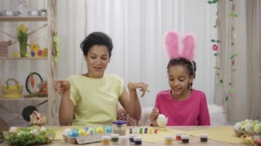 Komik tavşan kulaklı şirin küçük kız yumurtaları fırça ve boyalarla süslüyor. Afro-Amerikan anne ve kızı evde şen şakrak dekore edilmiş bir odada oturuyor. Mutlu Paskalyalar. Yavaş çekim hazır 59.97fps