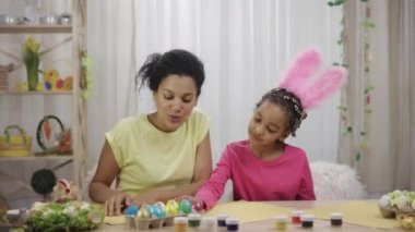 Komik tavşan kulaklı anne ve kızı boyalı yumurtaları düşünür. Afro-Amerikalı bir kadın ve küçük bir kız evdeki dekoratif bir odada masada oturuyorlar. Mutlu Paskalyalar. Yavaş çekim hazır 59.97fps.