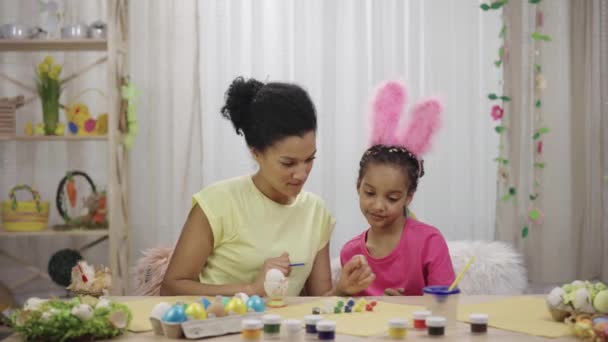 Mamãe ensina sua filha a pintar ovos com tintas e pincel. Mulher afro-americana e menina com orelhas de coelho estão sentados à mesa em quarto decorado. Feliz Páscoa. Movimento lento pronto 59.97fps. — Vídeo de Stock