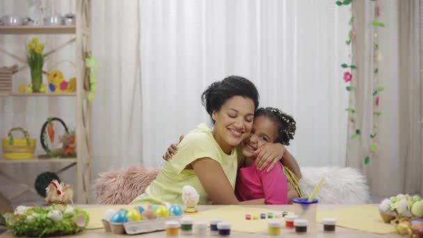 Mutter und Tochter liegen sich freudig in den Armen und unterhalten sich. Afroamerikanerin und kleines Mädchen sitzen zu Hause in einem festlich geschmückten Raum am Tisch. Frohe Ostern. Zeitlupe bereit 59.97fps. — Stockvideo