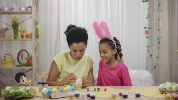 Mom teaches her daughter to paint eggs with paints and brush. African American woman and little girl with bunny ears are sitting at table in decorated room. Happy easter. Slow motion ready 59.97fps. — Stock Video