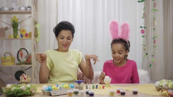 Roztomilá holčička s vtipnými králičími ušima zdobí vajíčka štětcem a barvami. Afroameričtí maminka a dcera sedí doma ve slavnostně zařízeném pokoji. Veselé Velikonoce. Zpomalení připraveno 59.97fps — Stock video