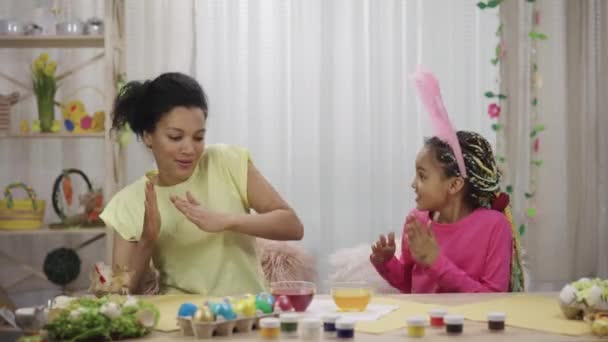 Komik tavşan kulaklı anne ve kızı komik dans ediyorlar. Afro-Amerikalı kadın ve küçük kız evdeki dekore edilmiş odada masada oturuyorlar. Mutlu Paskalyalar. Yavaş çekim hazır 59.97fps. — Stok video