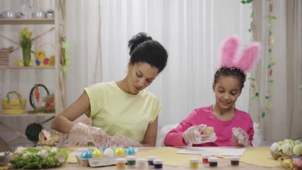 Mãe e filha com orelhas de coelho engraçado pintar ovos usando tinta amarela e rosa. Mulher afro-americana e menina estão sentados em quarto decorado em casa. Feliz Páscoa. Movimento lento pronto 59.97fps. — Vídeo de Stock