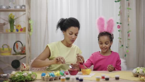 Mutter und Tochter mit lustigen Hasenohren tauchen Eier mit gelber und roter Farbe in Lebensmittelfarbe. Afroamerikanerin und kleines Mädchen sitzen in dekoriertem Raum. Frohe Ostern. Zeitlupe bereit 59.97fps — Stockvideo