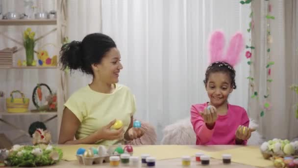 Komik tavşan kulaklı anne ve kızı renkli yumurtalarla gülüp eğleniyorlar. Afro-Amerikalı kadın ve küçük kız evdeki süslü odada masada oturuyorlar. Mutlu Paskalyalar. Yavaş çekim hazır 59.97fps — Stok video