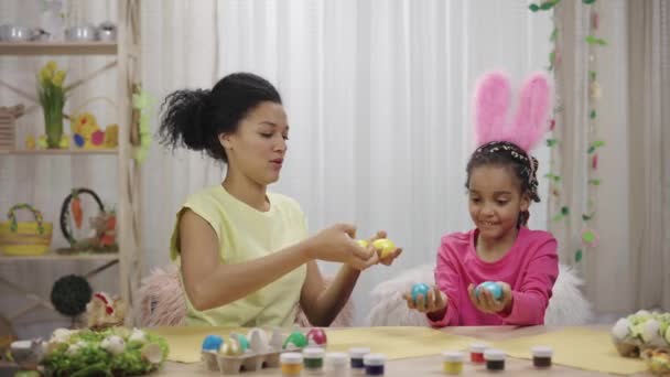 Mamá y su hija con divertidas orejas de conejo se ríen y se divierten con huevos de colores. Mujer afroamericana y niña pequeña sentadas en la mesa en la habitación decorada en casa. Feliz Pascua. Cámara lenta lista 59.97fps — Vídeos de Stock