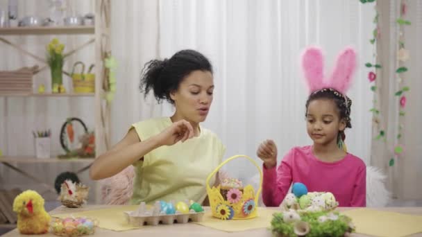 Mãe e filha com orelhas de coelho engraçado colocando bolo de Páscoa e ovos coloridos na cesta. Mulher afro-americana e menina sentada em quarto decorado. Feliz Páscoa. Movimento lento pronto 59.97fps. — Vídeo de Stock