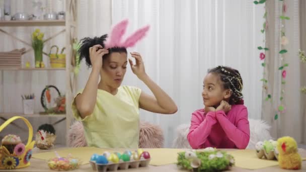 Mamãe tenta em sua filha engraçado orelhas de coelho rosa e ri alegremente. Mulher afro-americana e menina estão sentados à mesa em quarto festivamente decorado. Feliz Páscoa. Movimento lento pronto 59.97fps. — Vídeo de Stock