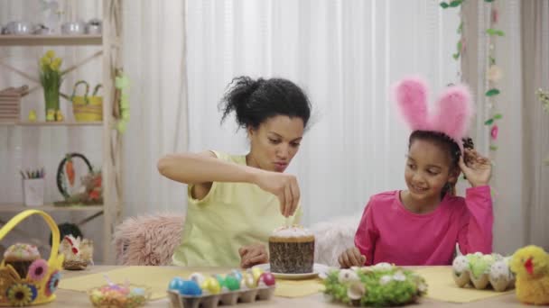 Mom and daughter with funny bunny ears decorate Easter cake with sweet candies. African American woman and little girl sittingin festively decorated room. Happy easter. Slow motion ready 59.97fps. — Stock Video