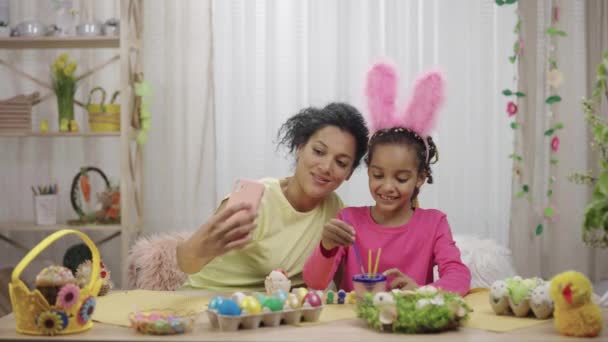 Mãe e filha com orelhas de coelho engraçado falar em uma chamada de vídeo usando o telefone. Mulher afro-americana e menina estão sentados à mesa em quarto decorado. Feliz Páscoa. Movimento lento pronto 59.97fps. — Vídeo de Stock