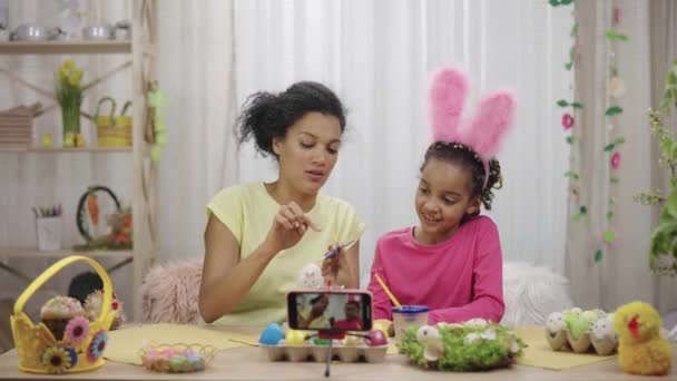 Mãe e filha com orelhas de coelho engraçado filmando no telefone eles pintando ovos. Mulher afro-americana e menina sentada em sala festivamente decorada. Feliz Páscoa. Movimento lento pronto 59.97fps. — Vídeo de Stock