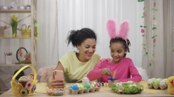 Maman et fille avec des oreilles de lapin drôles parlent sur un appel vidéo en utilisant le téléphone. Femme afro-américaine et petite fille sont assises à table dans une pièce décorée. Joyeuse Pâques. Ralenti prêt 59.97fps. — Video
