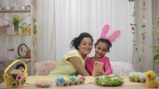 Ibu dan anak perempuan dengan telinga kelinci lucu mengambil selfie dan bersenang-senang. Wanita Afrika-Amerika dan gadis kecil duduk di meja di kamar berhias meriah. Selamat easter. Gerakan lambat siap 59.97fps. — Stok Video