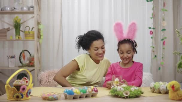 Mutter macht ihrer Tochter süßes Osterhuhn-Geschenk. Afroamerikanerin und kleines Mädchen sitzen zu Hause in einem festlich geschmückten Raum am Tisch. Frohe Ostern. Zeitlupe bereit 59.97fps. — Stockvideo