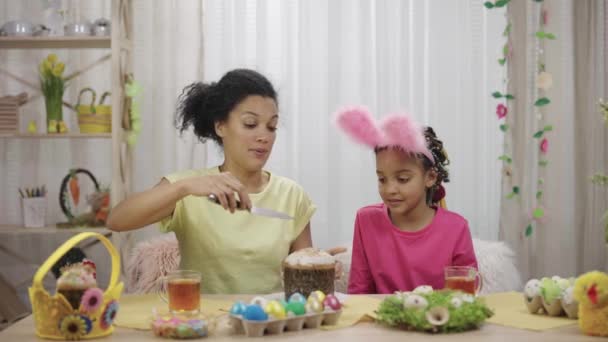 Mam snijdt een paastaart om haar dochter te behandelen. Afro-Amerikaanse vrouw en klein meisje zitten aan tafel in feestelijk ingerichte kamer thuis. Gelukkig Pasen. Slow motion klaar 59,97 fps. — Stockvideo