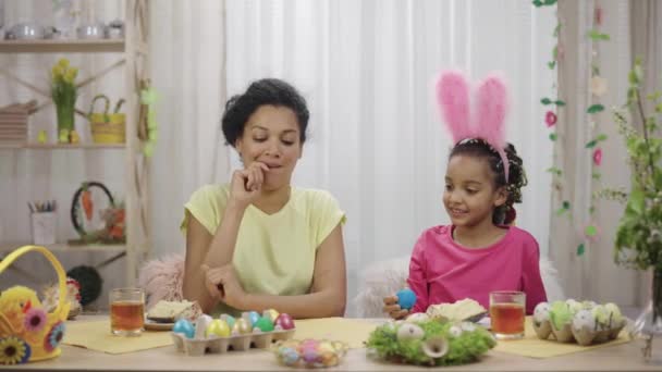 Anne ve kızı eğleniyor ve paskalya yumurtalarını kırıyor. Afro-Amerikalı kadın ve küçük kız evdeki dekore edilmiş odada masada oturuyorlar. Mutlu Paskalyalar. Yavaş çekim hazır 59.97fps. — Stok video