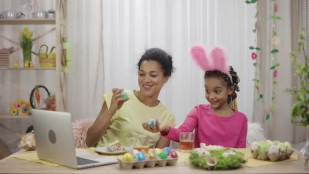 Mãe e filha conversam em videochamada via laptop, batem ovos e bebem chá com bolo de Páscoa. Mulher afro-americana e menina sentada em quarto decorado. Feliz Páscoa. Movimento lento pronto 59.97fps. — Vídeo de Stock