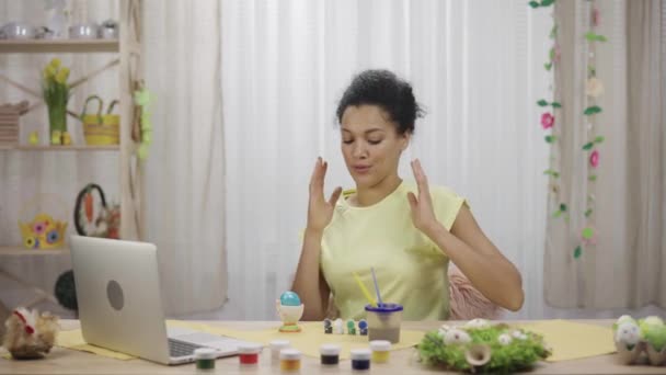 Cute African American woman talking by video call on laptop. A young woman teaches video lesson on painting Easter eggs, sitting at a decorated holiday table. Happy easter. Slow motion ready 59.97fps. — Stock Video