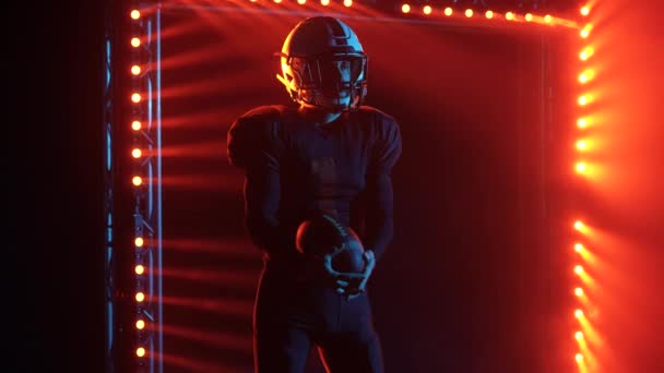 Portrét odhodlaného amerického fotbalisty držícího míč a připraveného ke hře. Muž pózující v tmavé aréně s červenými světly a kouřem. Důvěryhodný muž v uniformě a helmě. Zavřít. Zpomalený pohyb. — Stock video