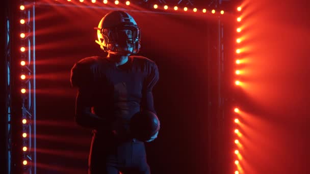 Retrato de determinado jugador de fútbol americano haciendo malabares con pelota y listo para el juego. Hombre posando en arena oscura con luces rojas y humo. Hombre confiado en uniforme y casco. De cerca. Movimiento lento. — Vídeos de Stock