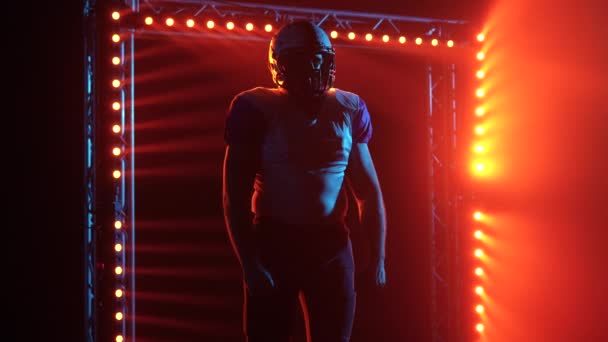 Silhouette af selvsikker quarterback kigge ind afstand. En amerikansk fodboldspiller i holduniform og hjelm står på stadion i mørke mod rødt baggrundslys. Filmisk slow motion-skud. Tæt på. – Stock-video