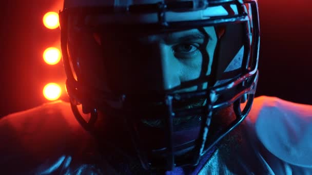 Giocatore professionista di football americano che guarda la telecamera. Uomo sicuro di sé in uniforme e casco in rosso blu retroilluminato stadio scuro. Sono pronto. Colpo al rallentatore cinematografico. Da vicino.. — Video Stock