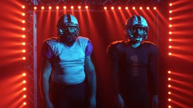 Spor kasklı ve üniformalı iki Amerikan futbolcu stadyuma girer. Kendine güvenen agresif oyuncular karanlıkta kırmızı ışığa karşı durarak rakiplerine meydan okuyorlar. Kapatın. Yavaş çekim.