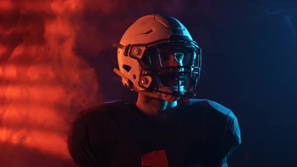 Retrato de quarterback mirando a su alrededor sobre fondo oscuro con luz azul roja y humo. Futbolista americano en uniforme de equipo y casco listo para jugar. Grabación cinematográfica en cámara lenta. De cerca.. — Vídeo de stock