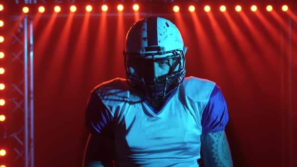 Retrato de quarterback confiante agressivo olhando para a câmera. Jogador de futebol americano em uniforme de equipe e capacete fica no estádio no escuro contra a luz vermelha. Fotografia em câmara lenta. Fechar — Vídeo de Stock