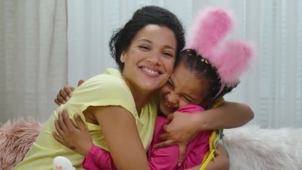 Mam en dochter met grappige konijnenoren die graag knuffelen en lachen. Afro-Amerikaanse vrouw en klein meisje zitten in versierde kamer thuis. Gelukkig Pasen. Langzame beweging. Sluiten.. — Stockvideo