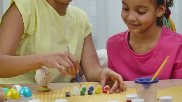 Mamma lär sin dotter att måla ägg med färg och pensel. Afroamerikansk kvinna och liten flicka med kaninöron sitter vid bordet i ett dekorerat rum. Glad påsk. Sakta i backarna. Närbild. — Stockvideo