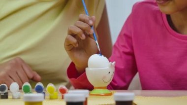 Komik tavşan kulaklı şirin küçük kız yumurtaları fırça ve boyalarla süslüyor. Afro-Amerikan anne ve kızı evde şen şakrak dekore edilmiş bir odada oturuyor. Mutlu Paskalyalar. Ağır çekim. Kapat..