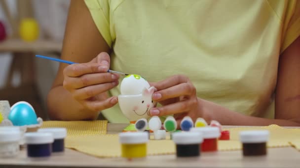 Mam leert haar dochter om eieren te schilderen met verf en borstel. Afro-Amerikaanse vrouw en klein meisje met konijnenoren zitten aan tafel in versierde kamer. Gelukkig Pasen. Langzame beweging. Sluiten.. — Stockvideo
