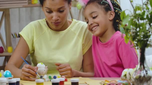 Mamá le enseña a su hija a pintar huevos con pinturas y pincel. Mujer afroamericana y niña con orejas de conejo están sentados en la mesa en la habitación decorada. Feliz Pascua. En cámara lenta. De cerca.. — Vídeos de Stock