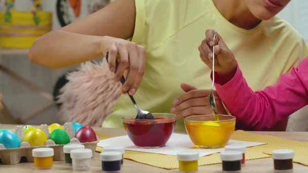 Mãe e filha com orelhas de coelho engraçado mergulha ovos em coloração de alimentos usando tinta amarela e vermelha. Mulher afro-americana e menina sentada em quarto decorado. Feliz Páscoa. Movimento lento. Fechar. — Vídeo de Stock