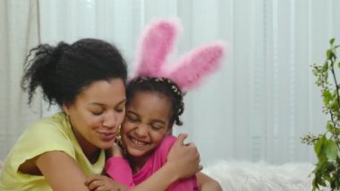 Komik tavşan kulaklı anne-kız kucaklaşıp gülümsüyor. Afrikalı Amerikalı bir kadın ve küçük bir kız evdeki süslü odada oturuyorlar. Mutlu Paskalyalar. Ağır çekim. Kapat..