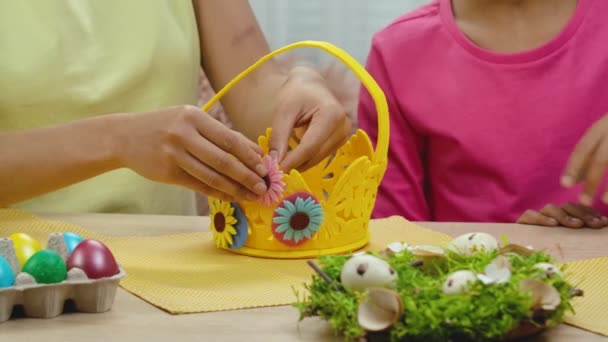 Mamma och dotter med roliga kaninöron dekorerar äggkorgen. Afroamerikansk kvinna och liten flicka sitter vid bordet i festligt inredda rum hemma. Glad påsk. Sakta i backarna. Närbild. — Stockvideo