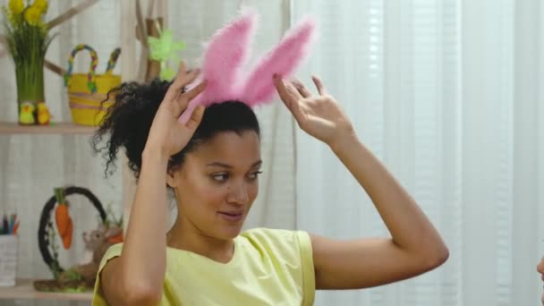 Maman essaie sur sa fille drôle oreilles de lapin rose et rit joyeusement. Femme afro-américaine et petite fille sont assises à table dans une pièce décorée de façon festive. Joyeuse Pâques. Au ralenti. Gros plan. — Video