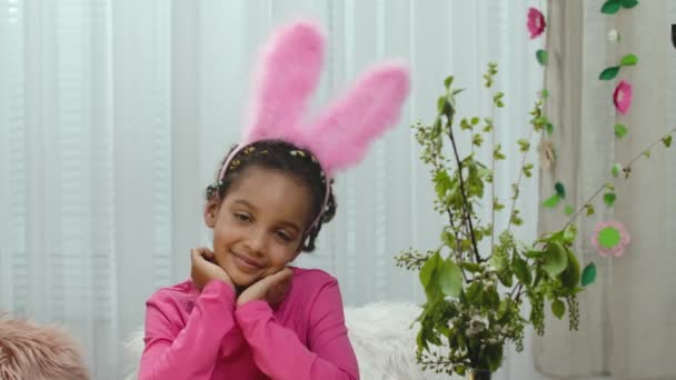 Una bambina carina guarda in divertenti orecchie di coniglio rosa guarda la fotocamera e sorride. Bambino afroamericano in posa sullo sfondo di una stanza luminosa. Buona Pasqua. Al rallentatore. Da vicino.. — Video Stock