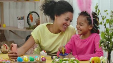 Komik tavşan kulaklı anne-kız telefonda video konuşmaları yapıyorlar. Afrikalı Amerikalı kadın ve küçük kız süslü bir odada masada oturuyorlar. Mutlu Paskalyalar. Ağır çekim. Kapat..