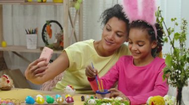 Komik tavşan kulaklı anne-kız telefonda video konuşmaları yapıyorlar. Afrikalı Amerikalı kadın ve küçük kız süslü bir odada masada oturuyorlar. Mutlu Paskalyalar. Ağır çekim. Kapat..